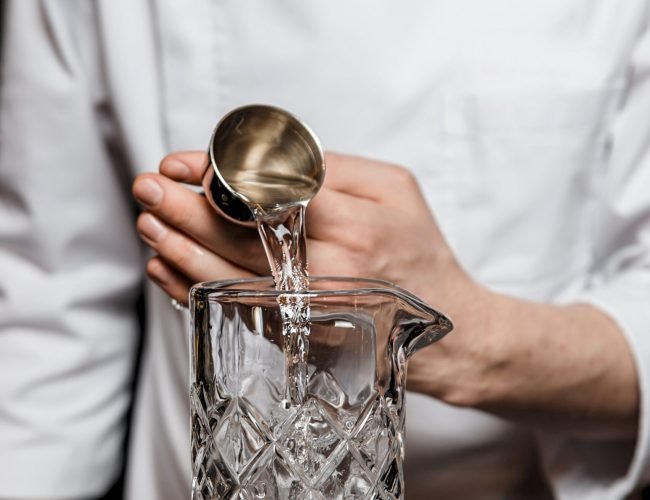 Bartender prepairing a cocktail at the bar, adding alcohol to the mixing glass using a jigger